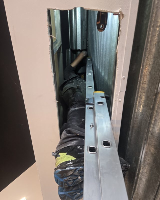 Technicien JBE Automatisme allongé dans le faux plafond, jambes dépassant de la trappe, en train de remplacer le moteur central d'un rideau métallique