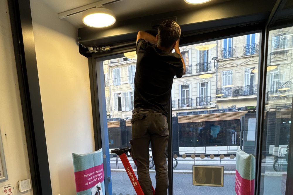 Technicien JBE Automatisme sur un escabeau, intervenant sur l'opérateur d'une porte automatique coulissante à l'entrée d'un magasin