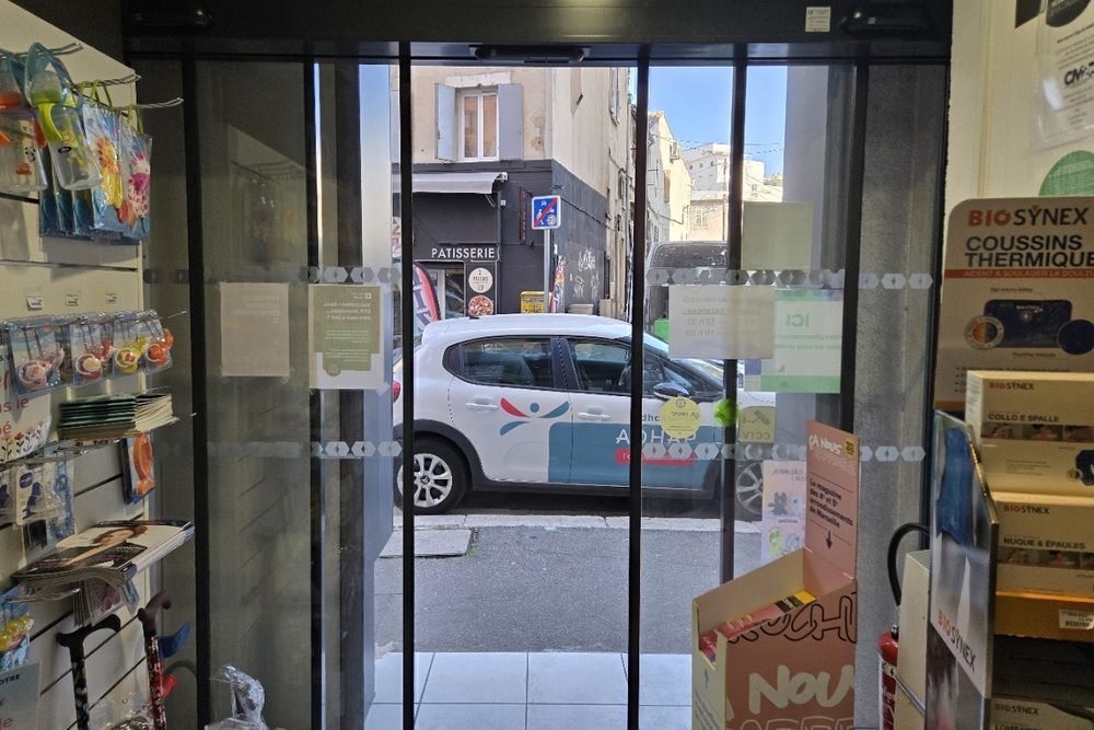 Porte coulissante automatique ouverte à l'entrée d'une pharmacie marseillaise, vue depuis l'intérieur, rue et immeubles au second plan