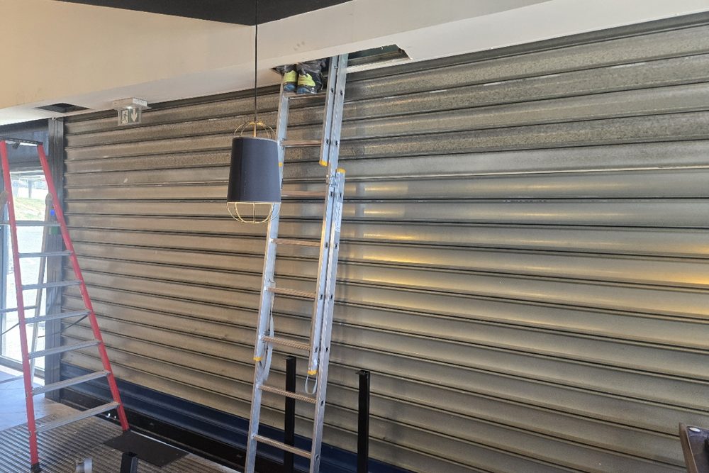 Intérieur d'un commerce, rideau métallique fermé, échelle dressée vers une trappe du faux plafond où intervient un technicien